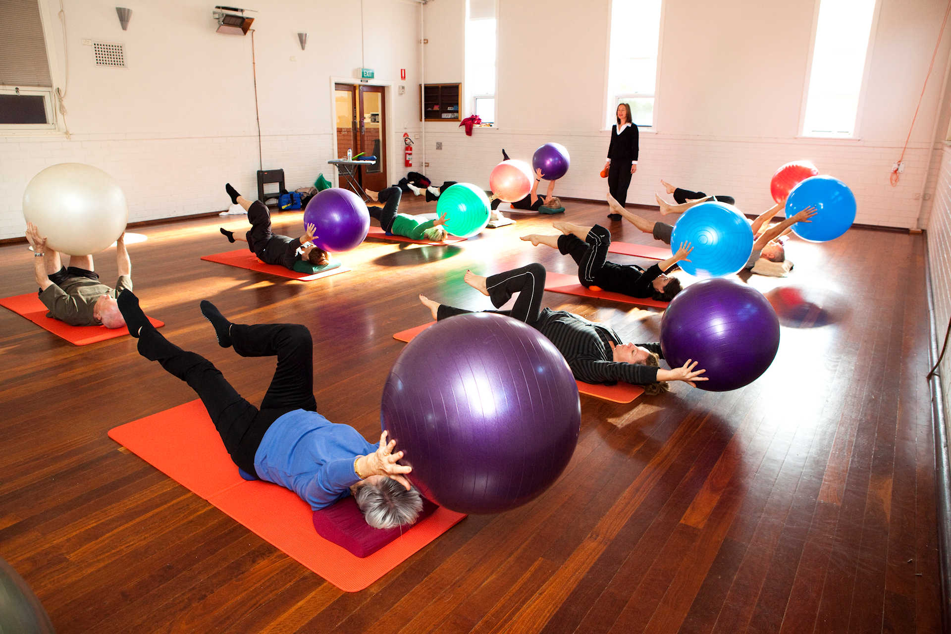 pilates on the ball group class in a hall lying on their back doing the scissors exercise whilst holding the ball above their heads with physio Jodie Krantz instructing.