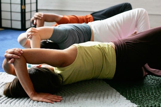 three people lying on their side on the floor doing a feldenkrais awareness through movement lesson, elbow over the head hand on floor other hand holding elbow, legs in open book position